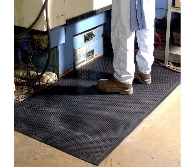Black Safety Scrape nitrile rubber mat on a workshop floor beside a machine, with a worker standing on it