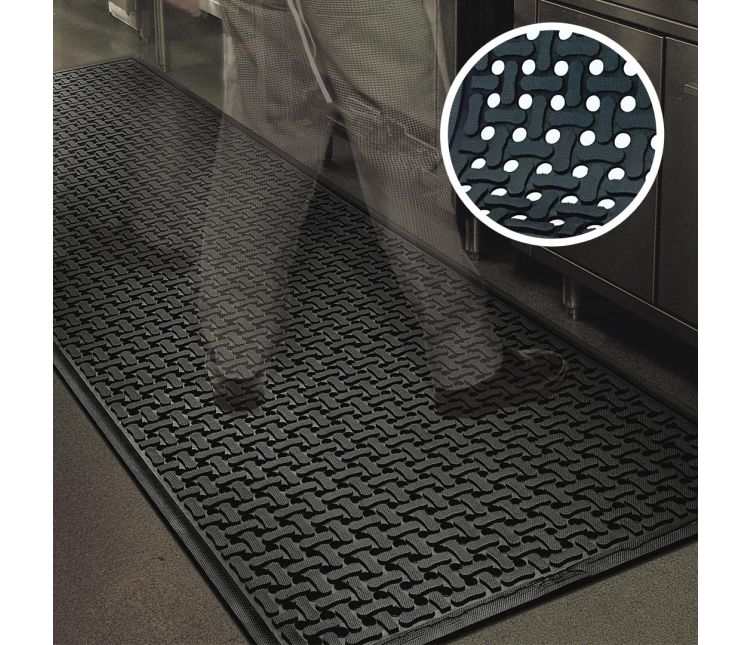 Black Comfort Flow drainage-hole rubber mat on a commercial kitchen floor beside stainless steel units, with a close-up inset of the flow-through holes