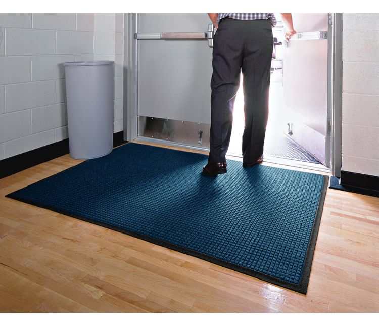 Blue WaterHog Classic entrance mat with a black rubber border on a wooden floor inside a commercial double doorway, as a person steps out through the open door