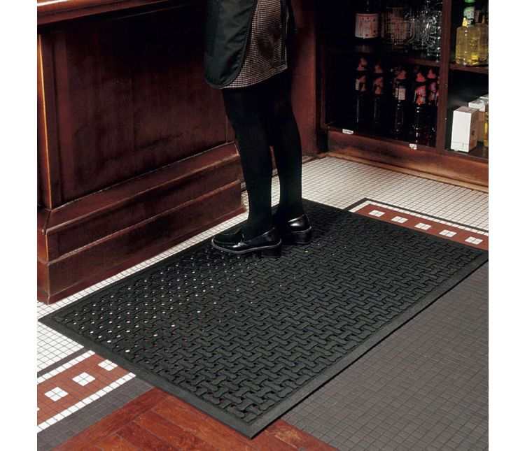 Black Comfort Flow drainage-hole rubber mat on a tiled floor behind a bar, with a member of staff standing on it in black shoes