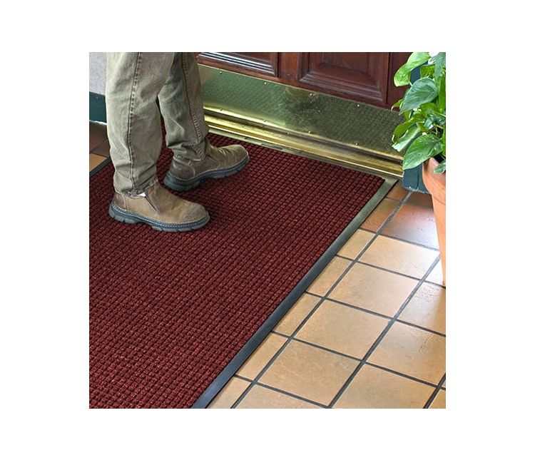 Person in work boots standing on a red WaterHog Classic entrance mat inside a wooden door with a brass kick plate, on a tiled floor